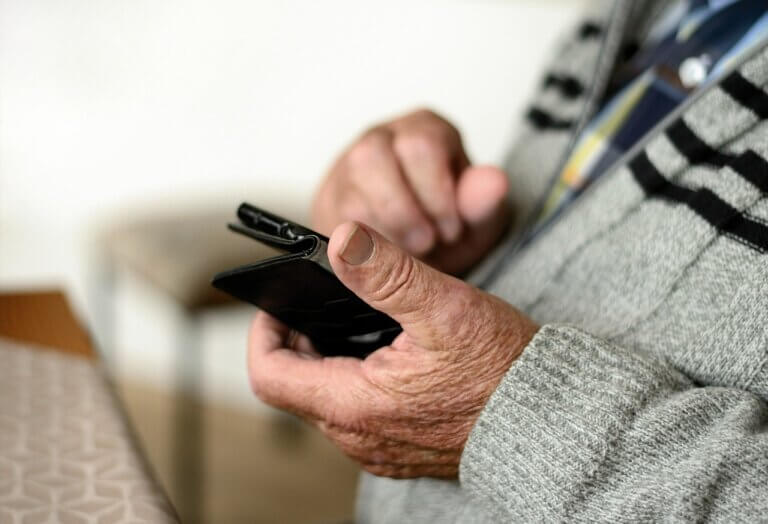A person uses a mobile device, their hands show signs of advanced age.